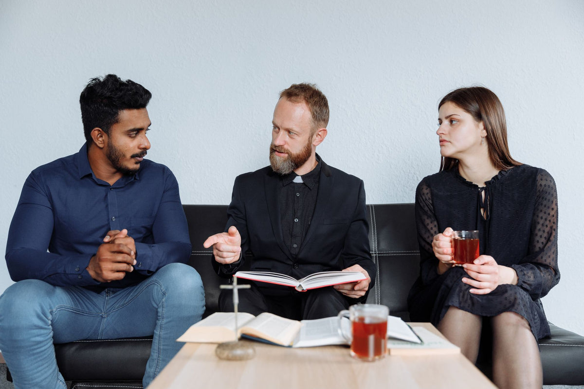 A priest and two young Catholics talking over tea and an open Bible